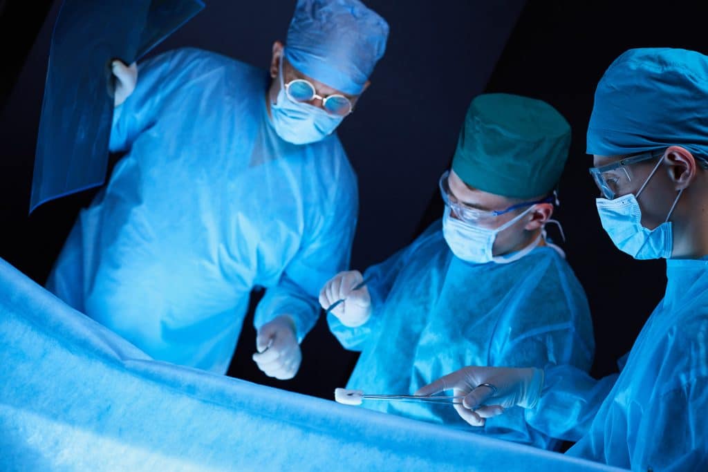 Three surgeons in medical scrubs and masks perform surgery under bright lights, with one holding an X-ray and others using surgical instruments over a patient.