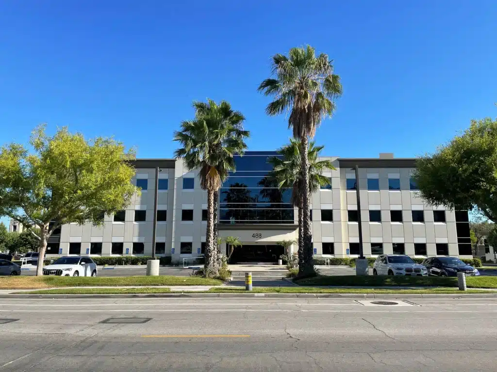 A three-story office building with reflective windows, palm trees in front, and parked cars along the entrance under a clear blue sky.