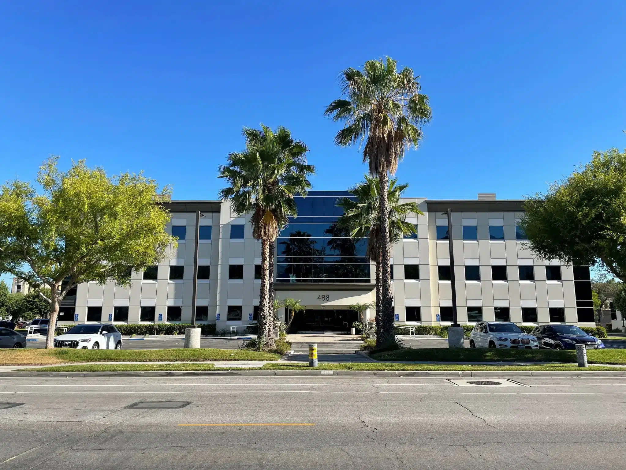 A three-story office building with reflective windows, palm trees in front, and parked cars along the entrance under a clear blue sky.