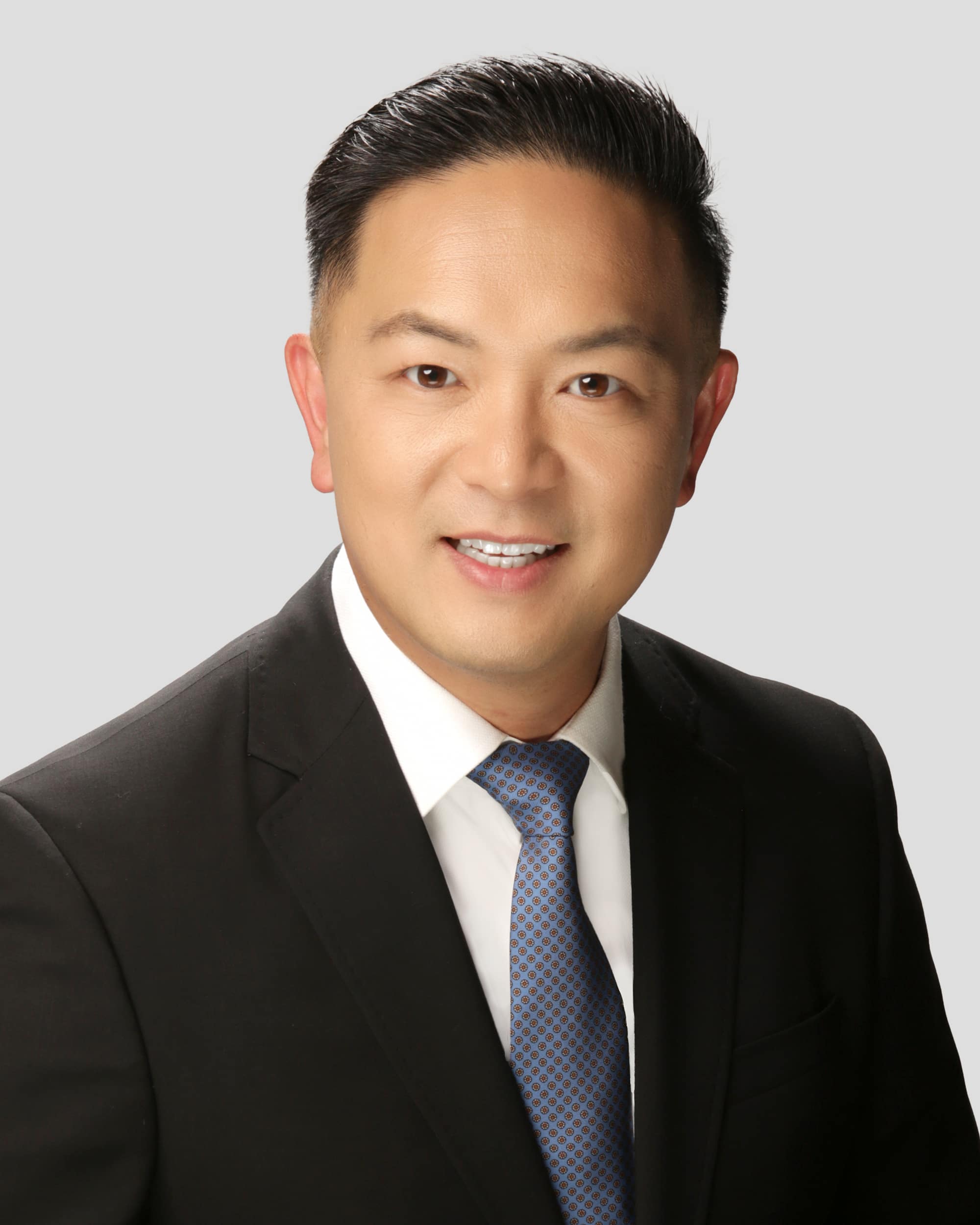 A man in a black suit, white shirt, and blue patterned tie smiles at the camera against a plain light gray background.