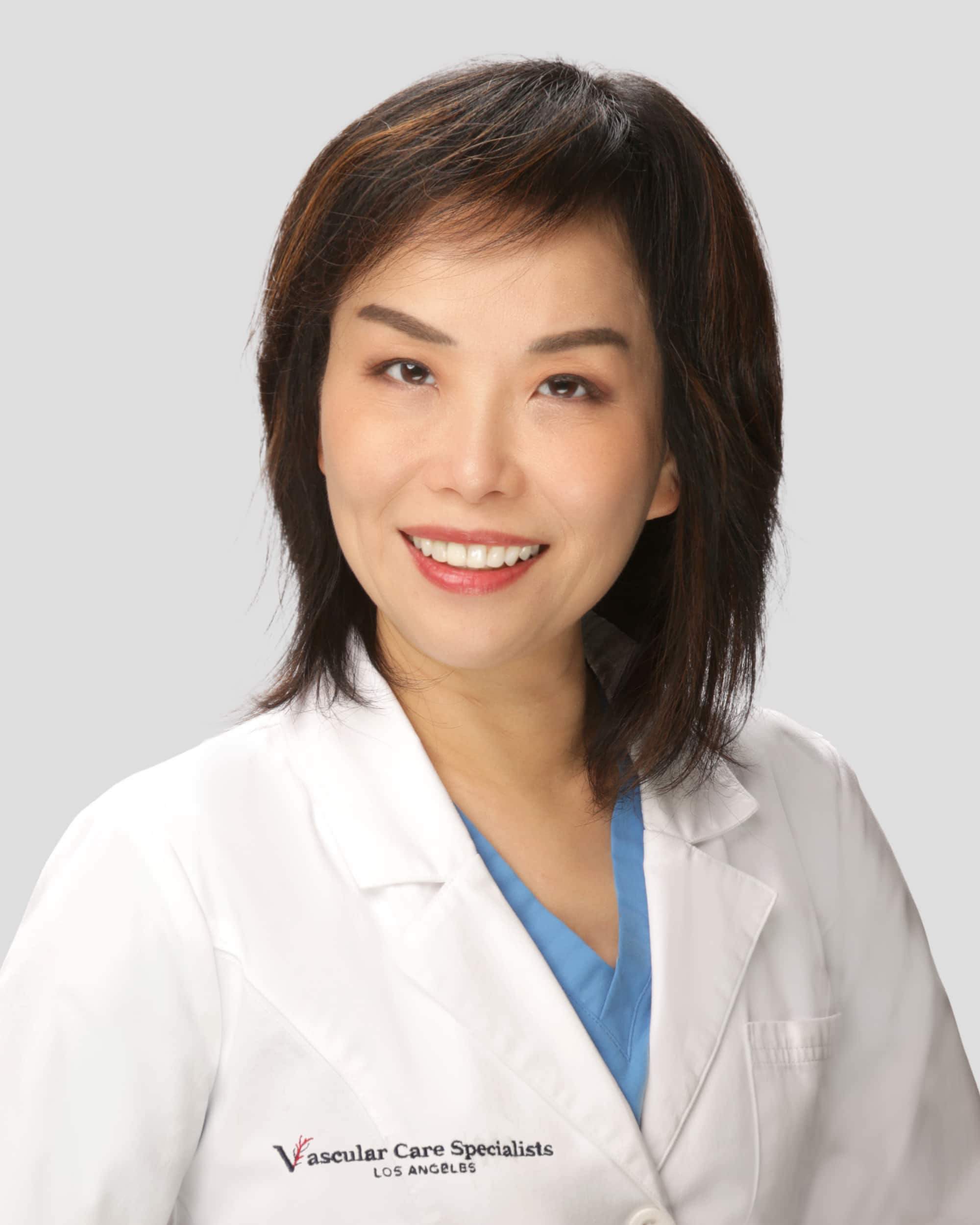 A woman in a white lab coat and blue scrubs smiles at the camera against a plain background. The coat reads "Vascular Care Specialists Los Angeles.