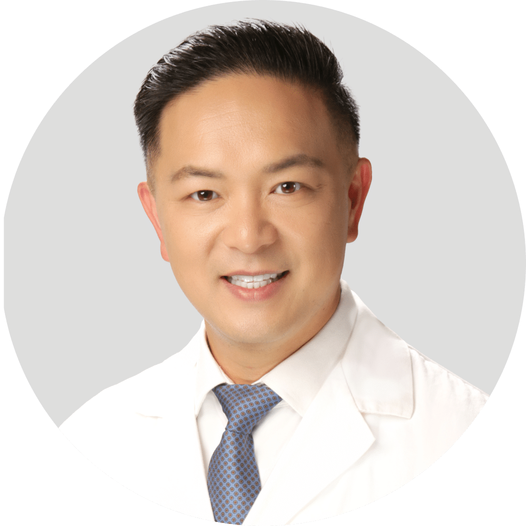 A man wearing a white lab coat and blue tie smiles at the camera against a light gray background.