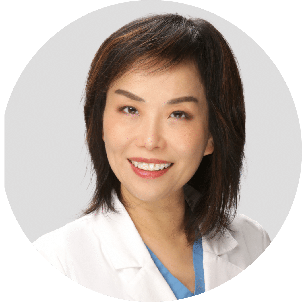 A woman with straight dark hair wearing a white lab coat and blue top, smiling at the camera against a plain light background.