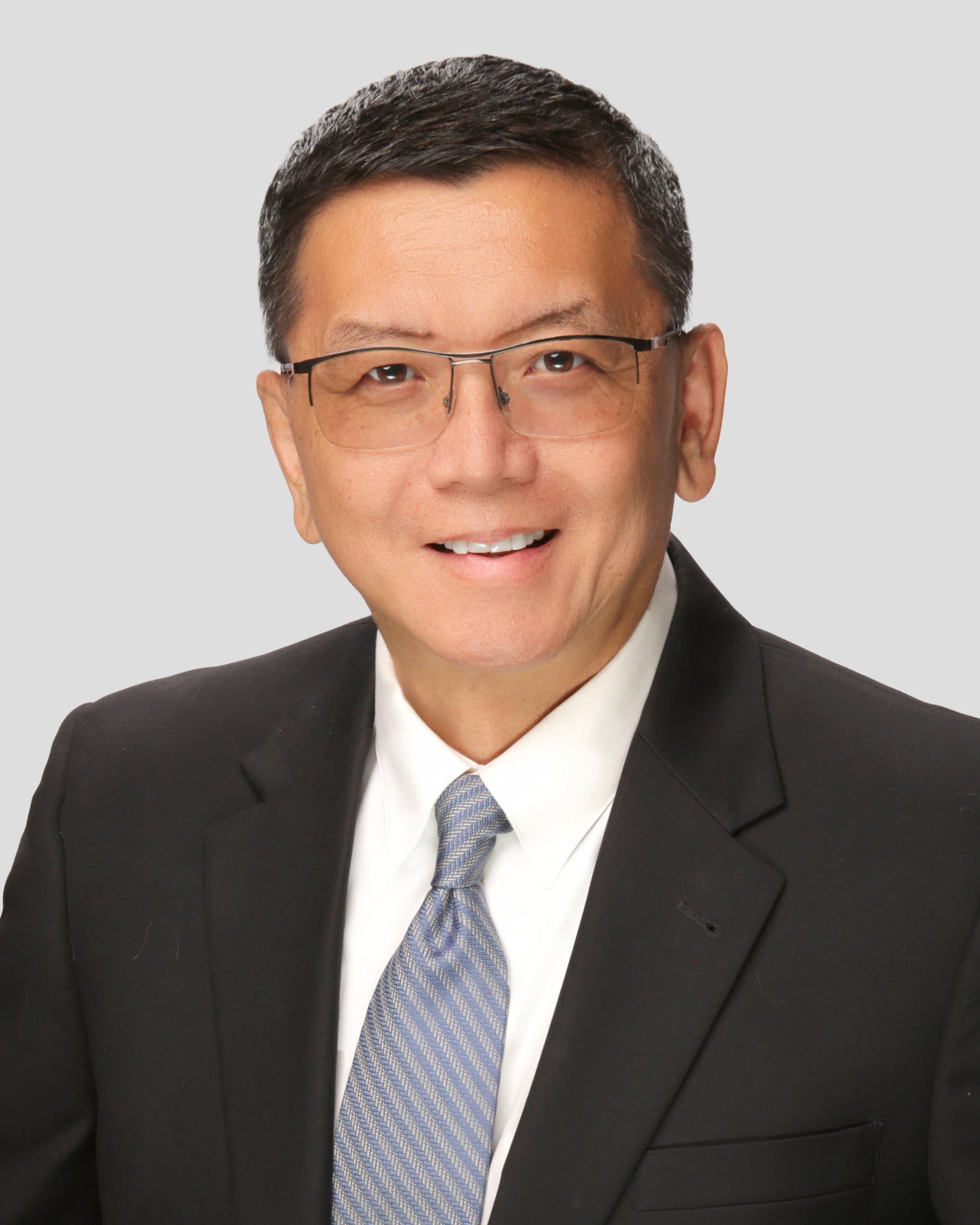 A man wearing glasses, a dark suit, white shirt, and striped tie poses for a professional headshot against a plain light gray background.