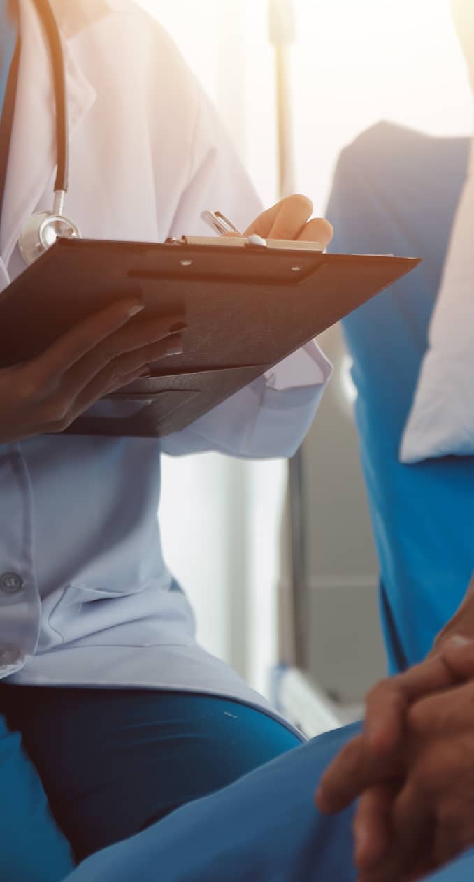 A doctor holding a clipboard and taking notes while sitting next to a patient in a hospital setting.
