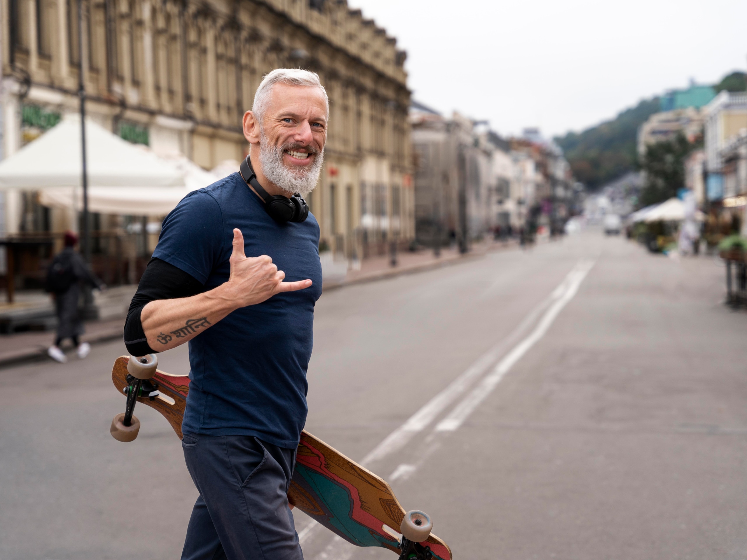 A man with grey hair and beard smiles and holds a skateboard while making a shaka sign, standing on an empty city street.