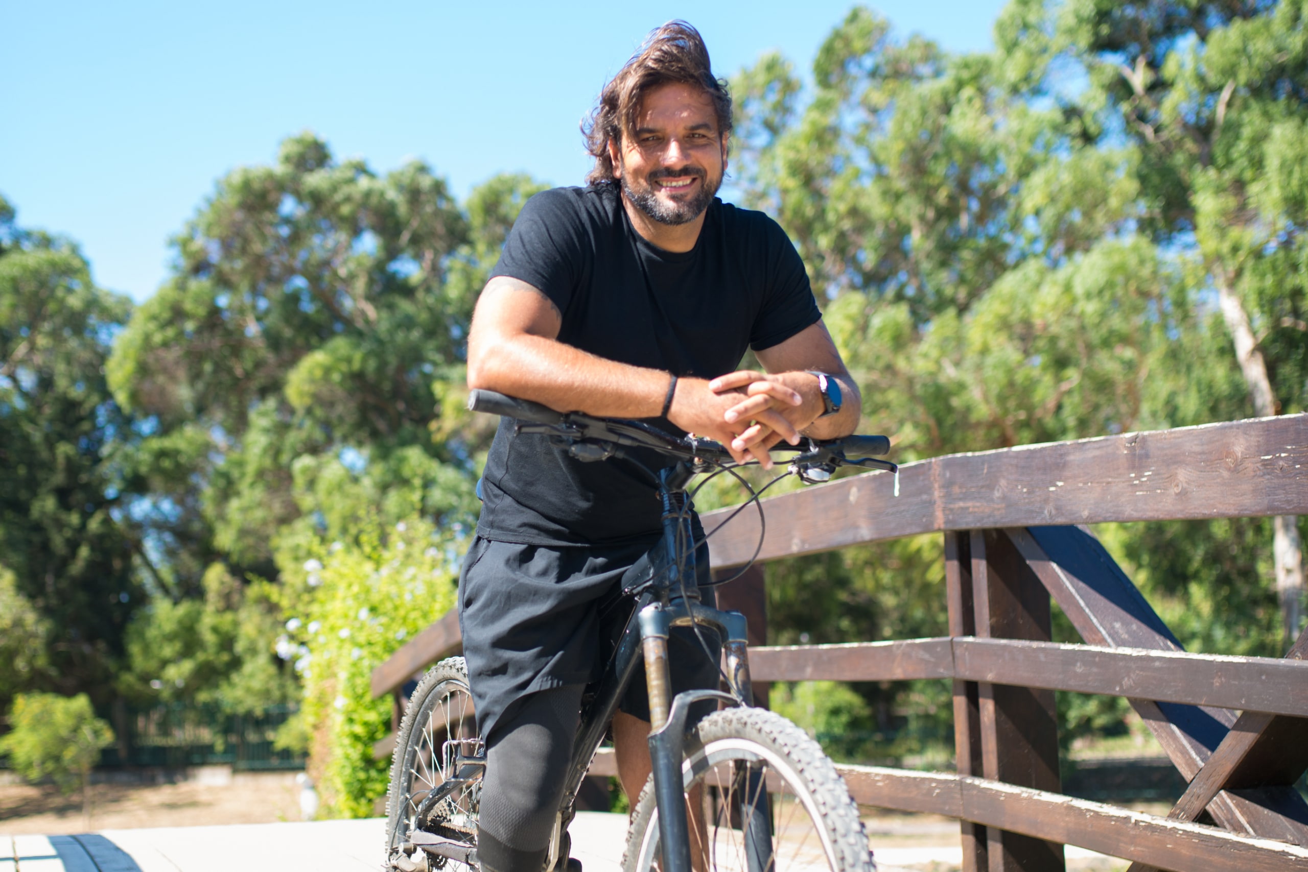 A man with a prosthetic leg stands outdoors, leaning on a bicycle and smiling, with trees and a wooden fence in the background.