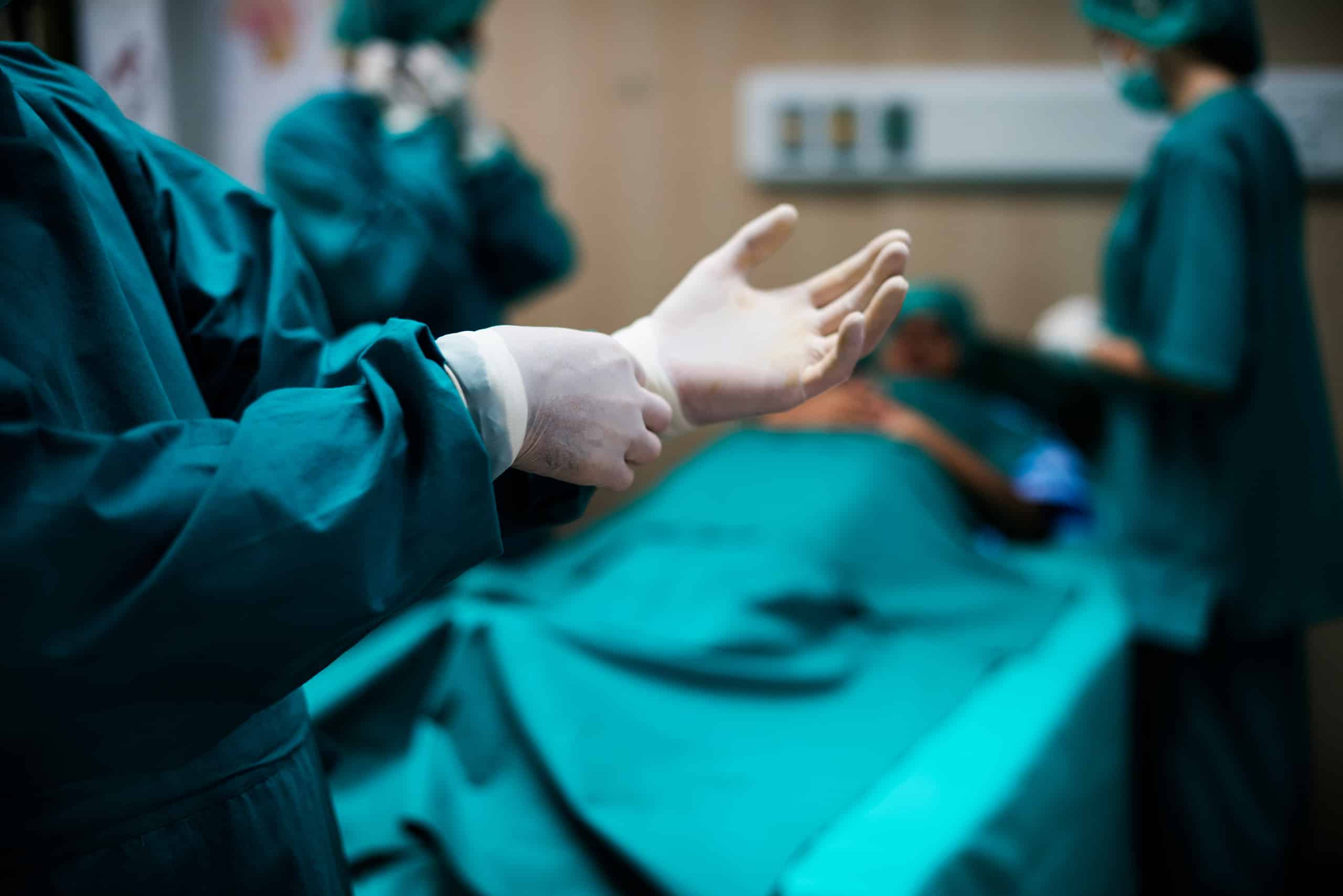 A surgeon puts on sterile gloves in an operating room, with medical staff and a patient on the operating table in the background.