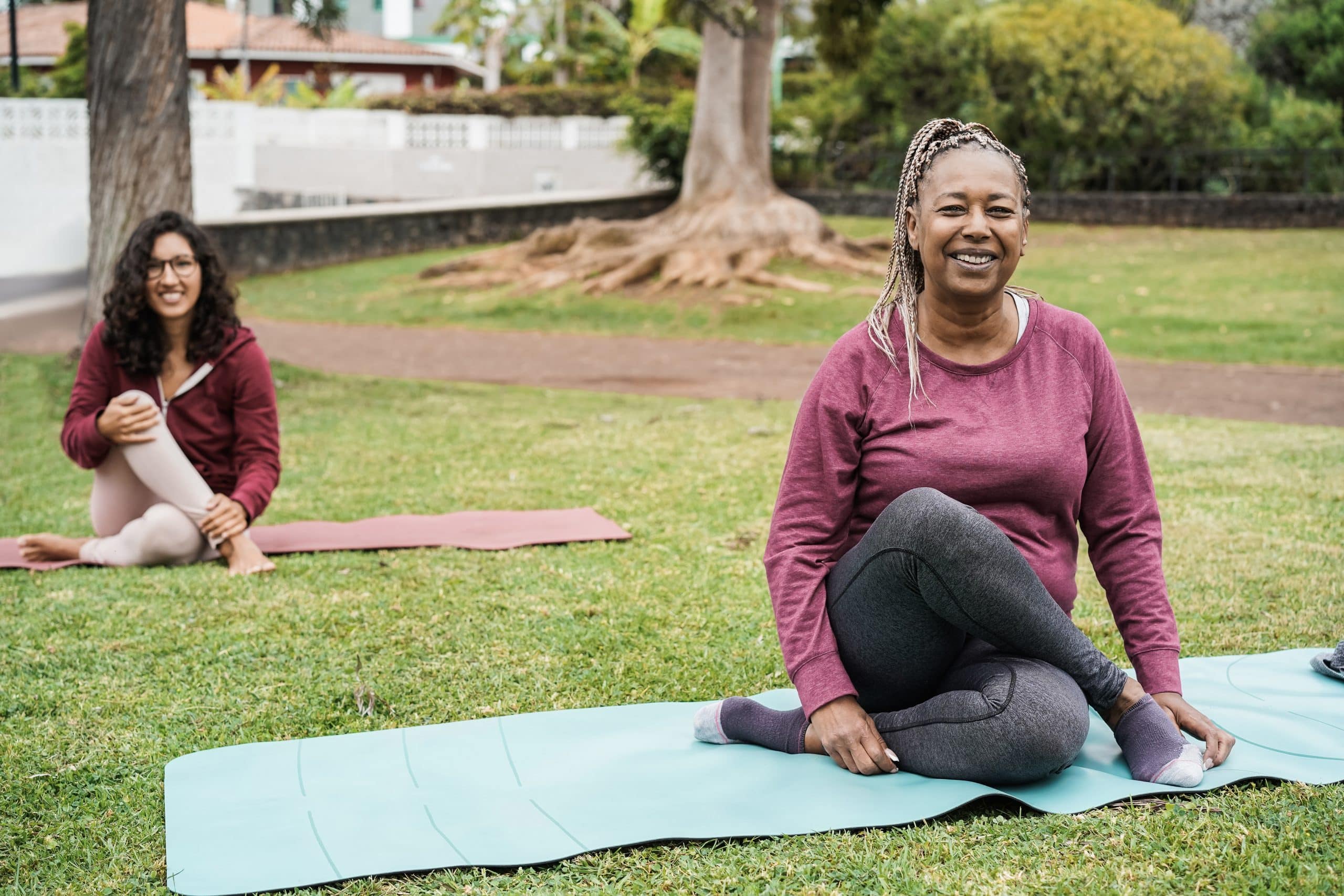 Two women sit on yoga mats outdoors, stretching and smiling during a yoga session in a grassy park.