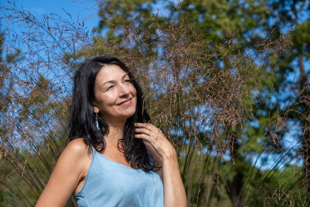 A woman with long dark hair, wearing a light blue sleeveless top, stands outdoors among tall grasses, smiling and looking slightly to the side. Trees and blue sky are in the background.