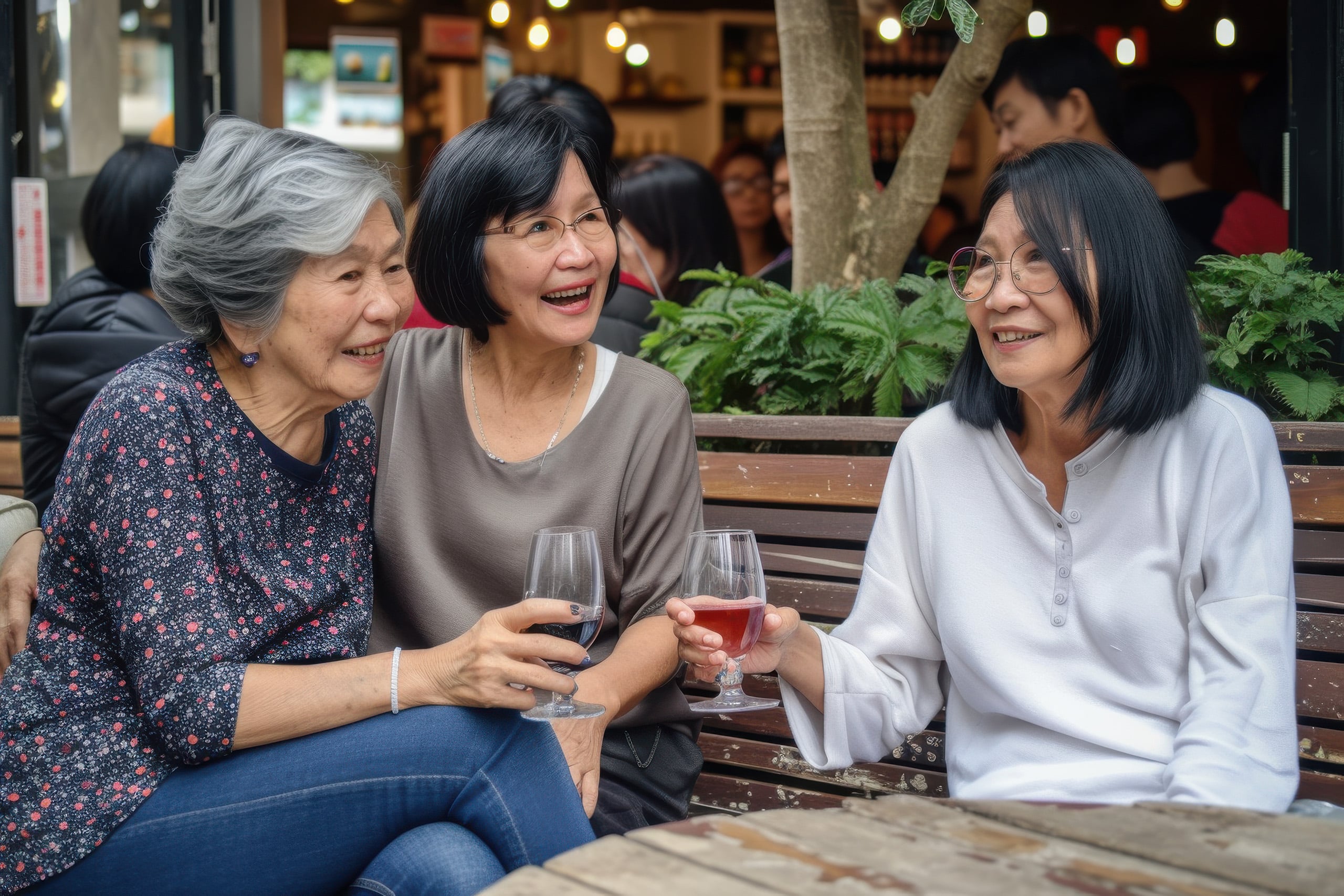 Three older women sit on a bench outdoors, holding wine glasses and smiling while having a conversation. There are plants and other people in the background.