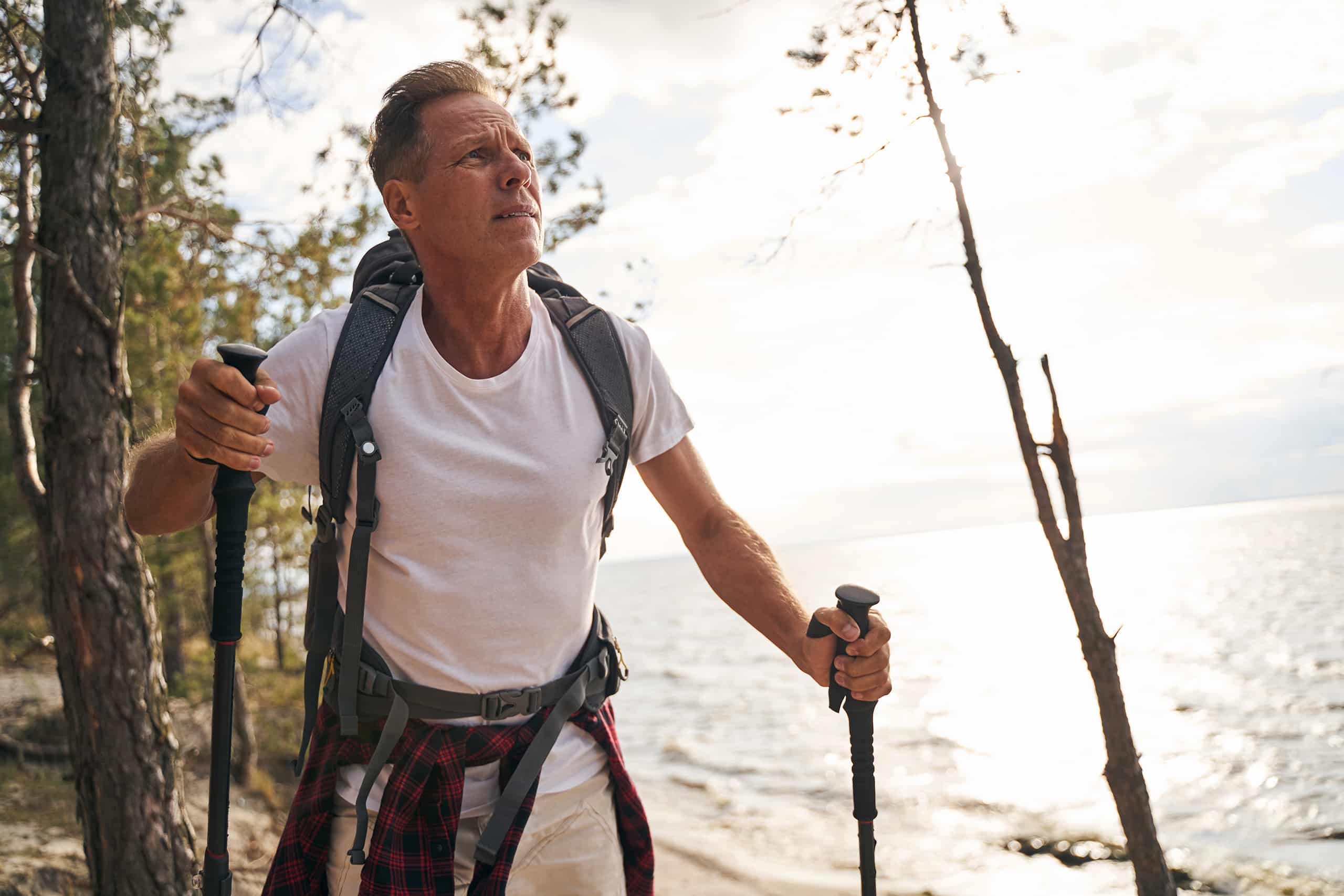 Man in a white t-shirt and backpack hiking with trekking poles along a wooded lakeside trail, sunlight reflecting off the water in the background.