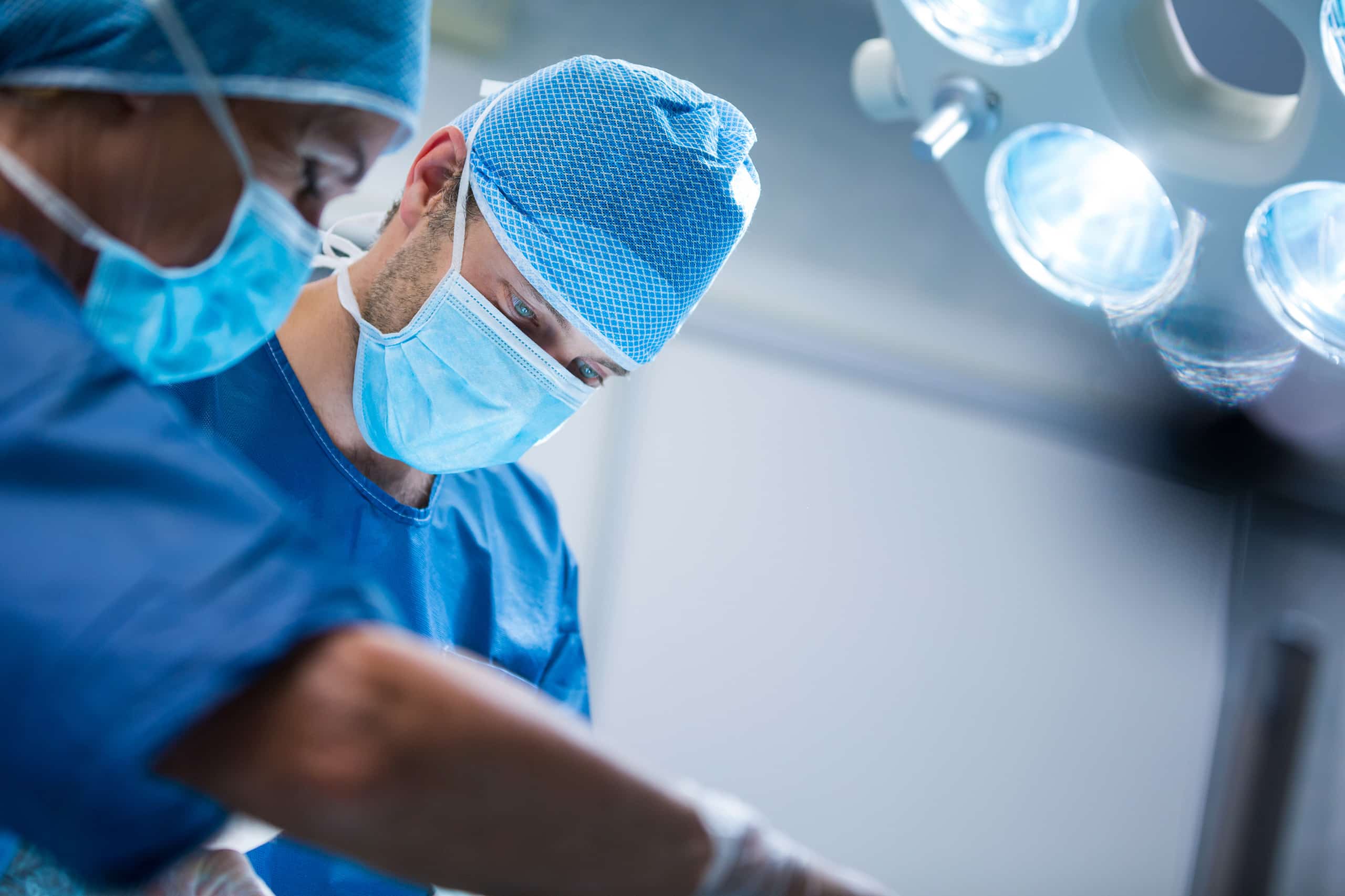 Two surgeons wearing blue scrubs, masks, and caps operate under bright surgical lights in an operating room.
