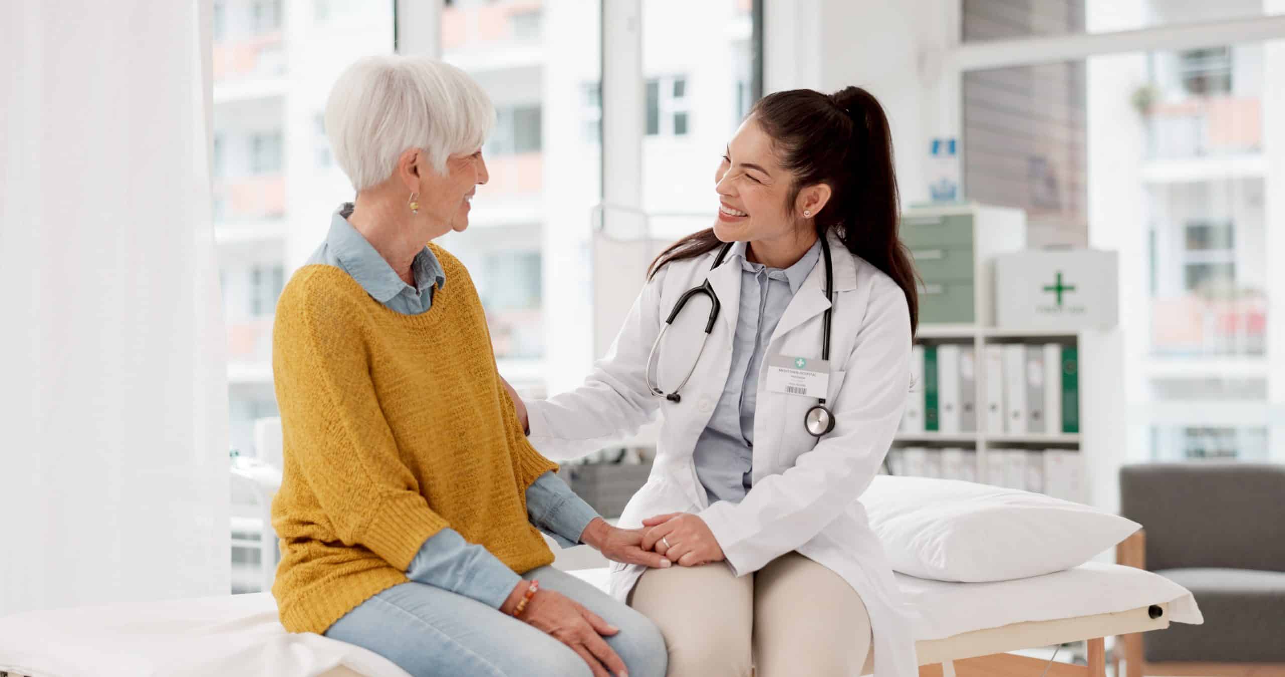 A doctor with a stethoscope talks to and comforts an older woman in a medical office.