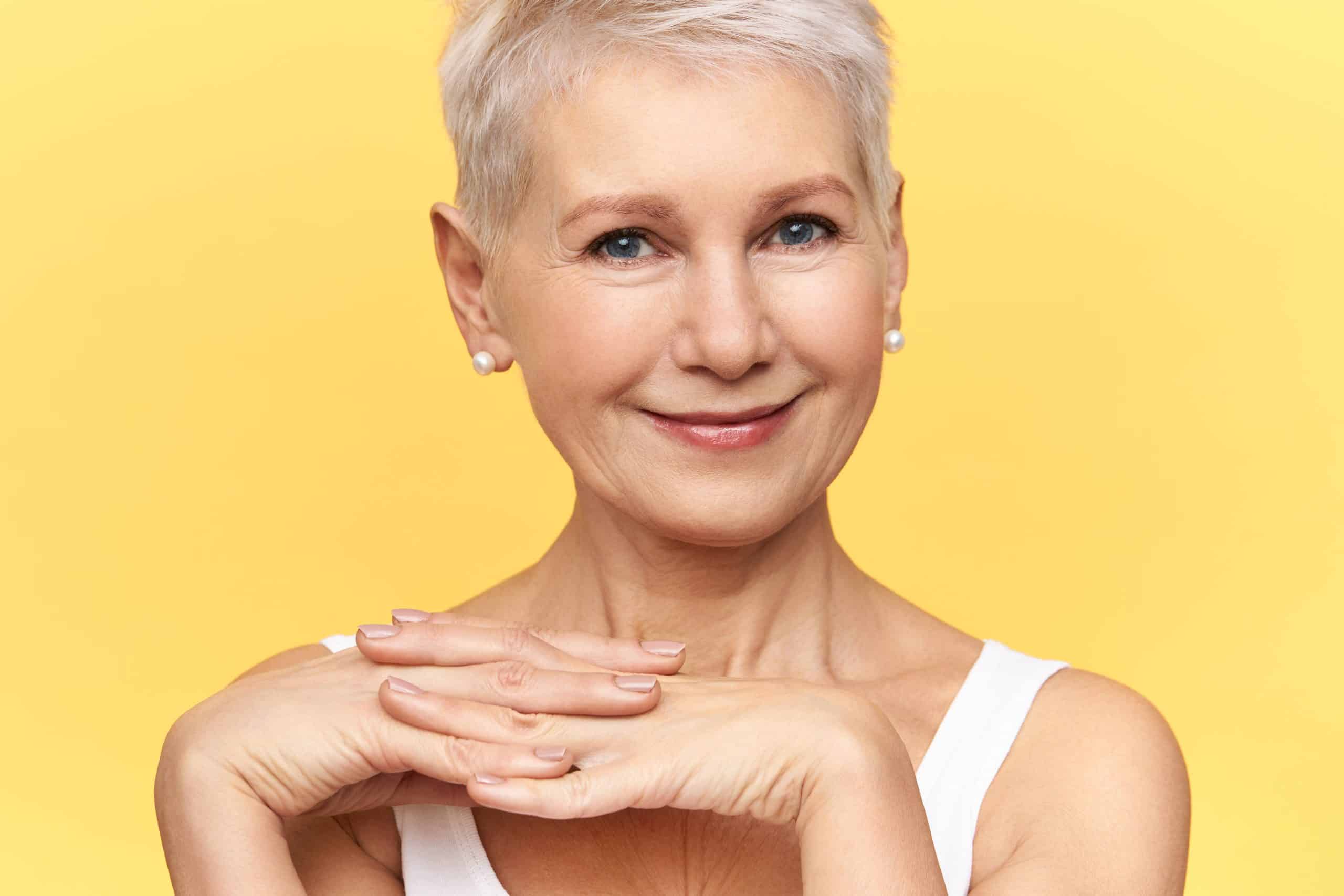 Older woman with short white hair and pearl earrings smiles gently, resting her chin on her hands, against a plain yellow background.