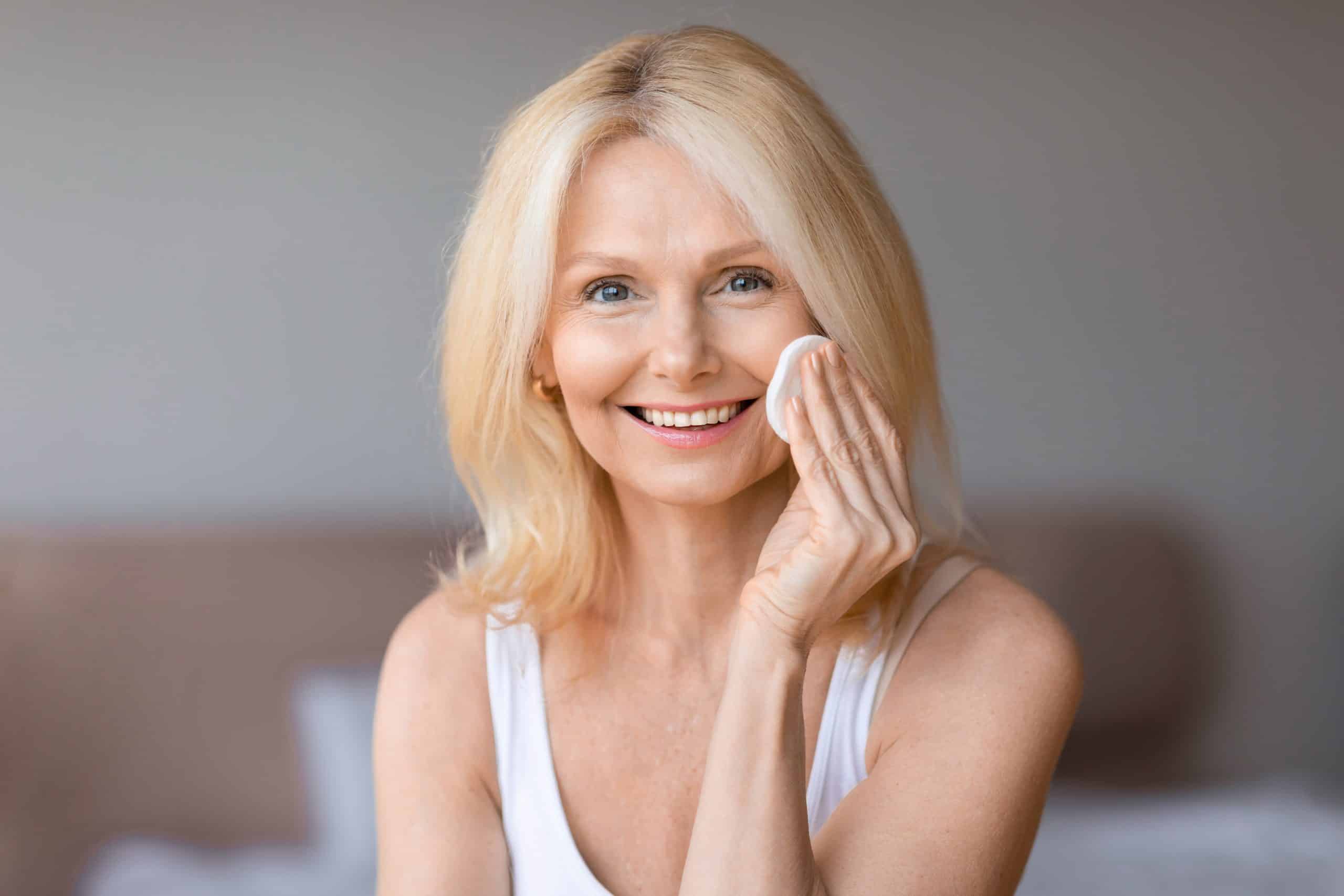 Smiling woman with blonde hair uses a cotton pad on her cheek, wearing a white tank top in a softly lit room.