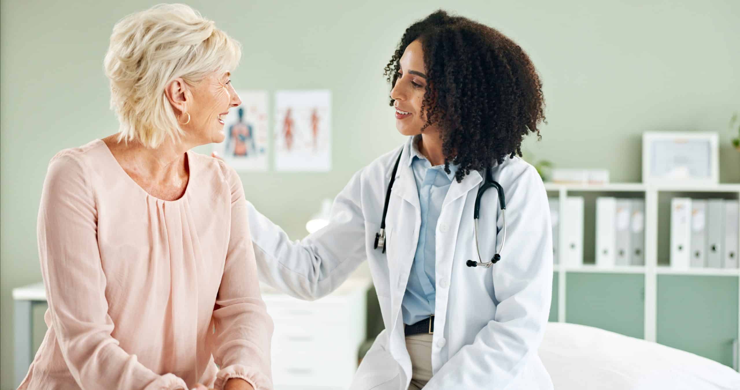 A doctor with a stethoscope talks to an older woman, sitting together in a medical office. The doctor is smiling and gently touching the woman’s shoulder.