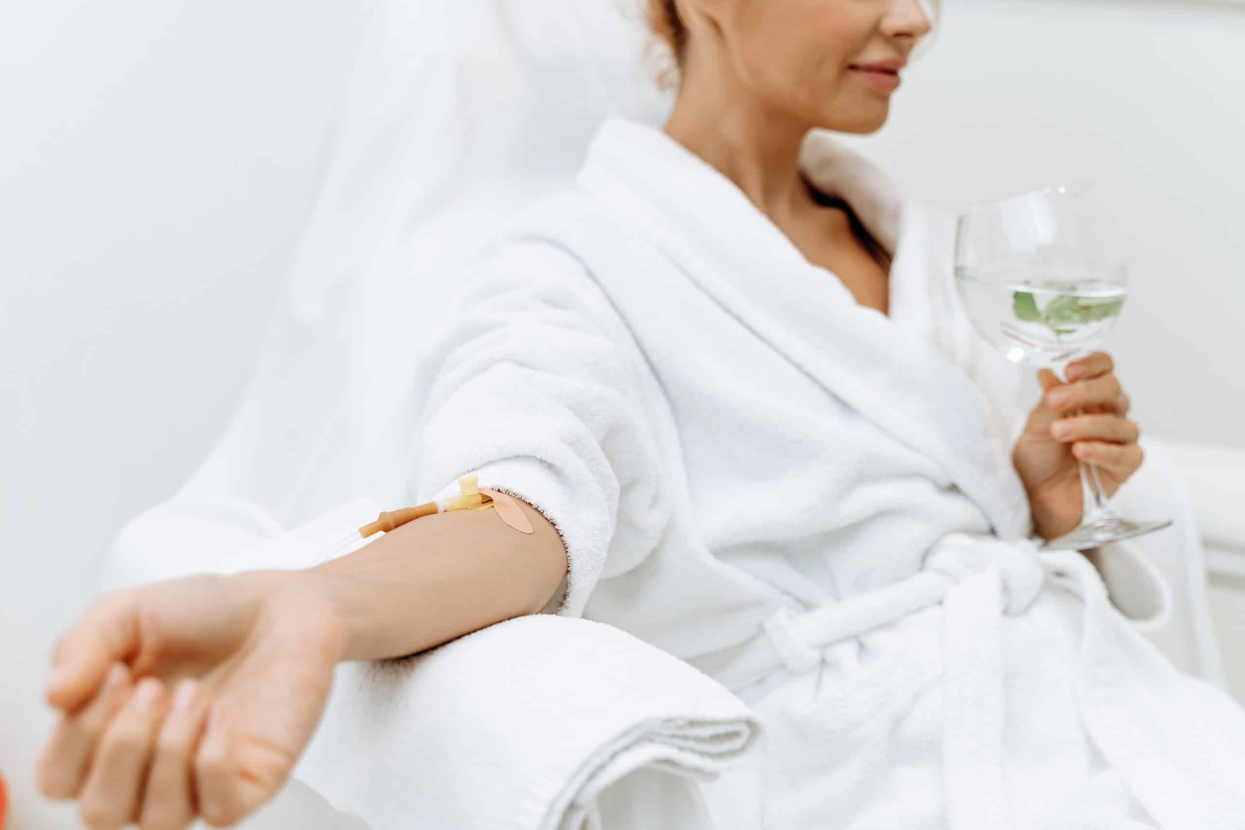 A woman in a white robe sits in a chair with an IV in her arm, holding a glass of water with a mint leaf.