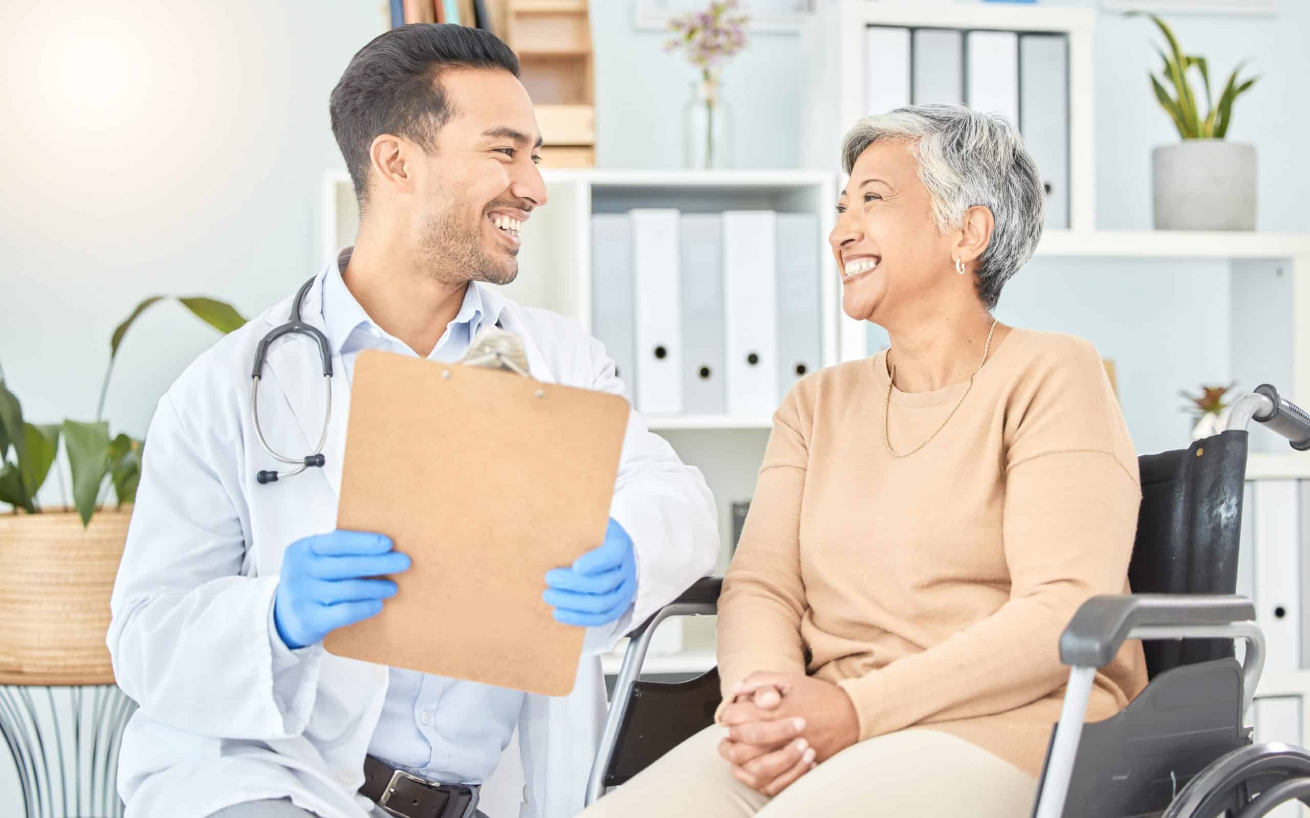 A doctor holding a clipboard smiles and talks with a woman in a wheelchair; both appear to be in a medical office setting.