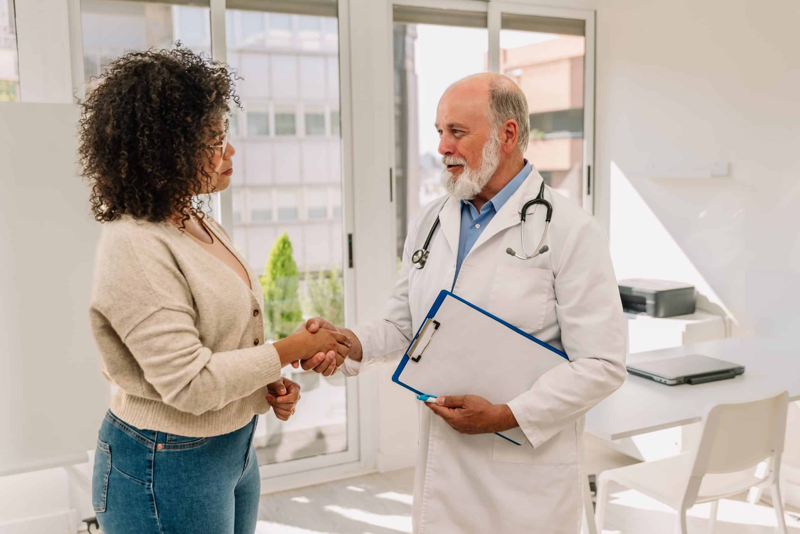 A doctor holding a clipboard shakes hands with a woman in an office setting, both standing and facing each other.