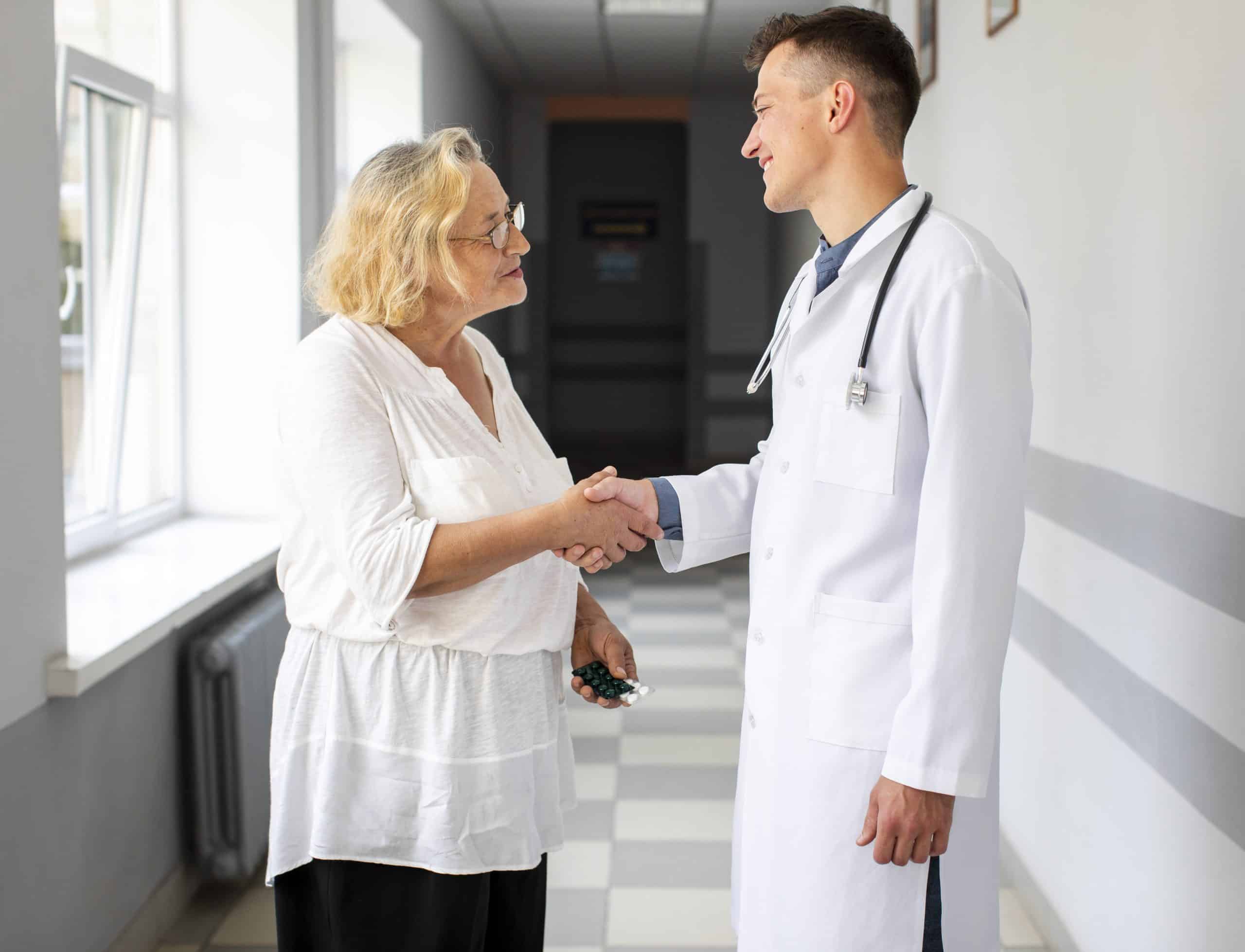 An older woman shakes hands with a male doctor in a hallway, both smiling and making eye contact.
