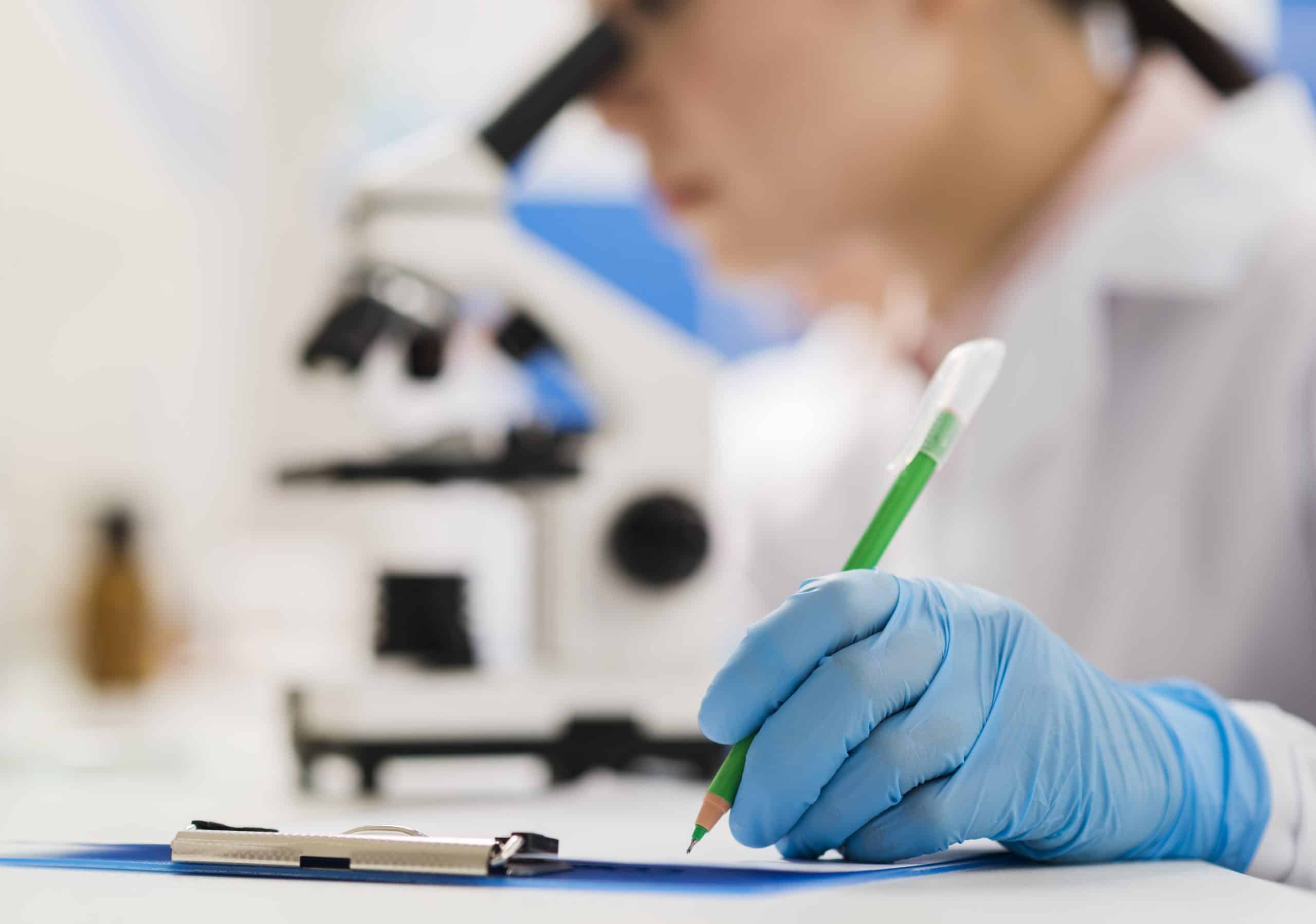 A person wearing blue gloves writes in a notebook with a green pencil near a clipboard, with a microscope visible in the background.