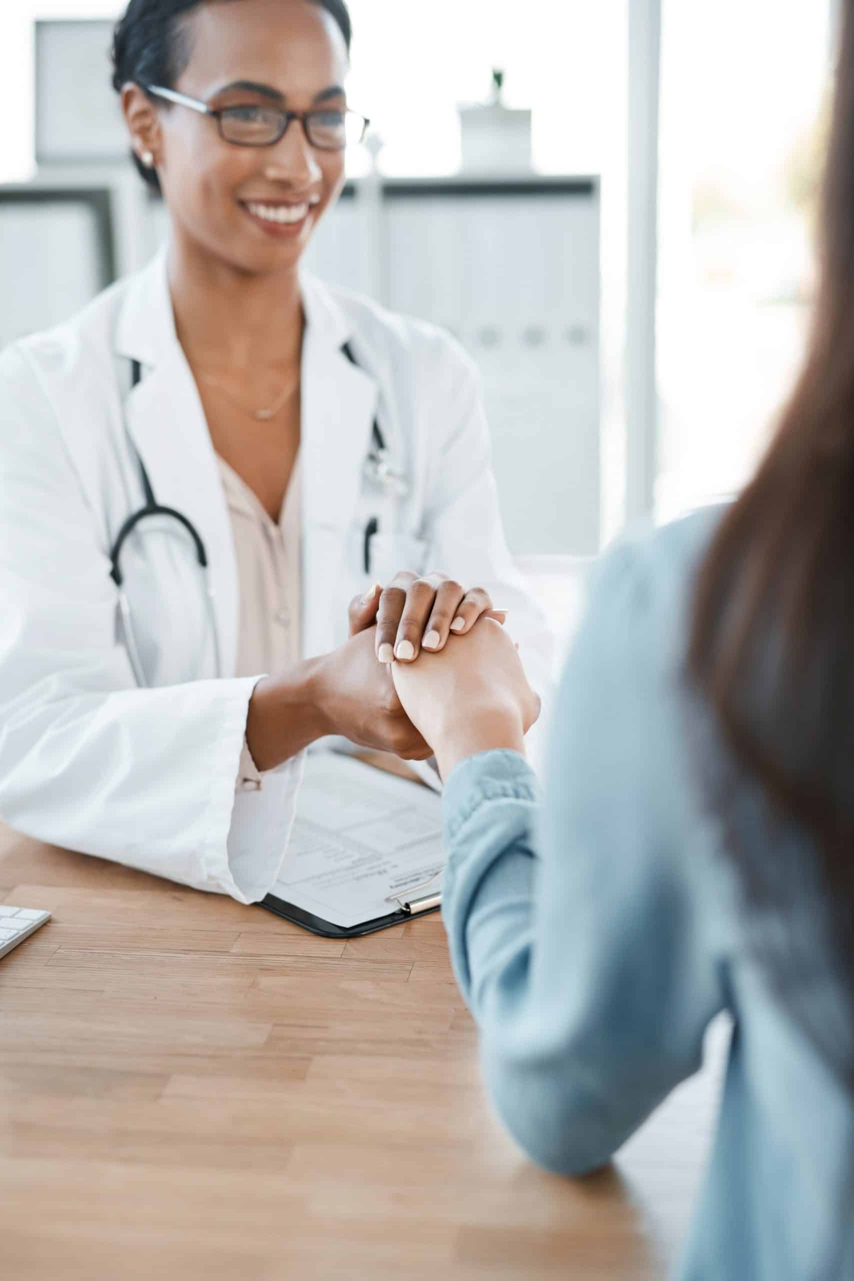 A doctor in a white coat holds a patient's hand across a desk, suggesting comfort and support during a medical consultation.