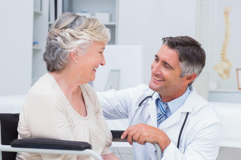 A senior woman in a wheelchair smiles while talking to a male doctor in a white coat and stethoscope in a medical office.