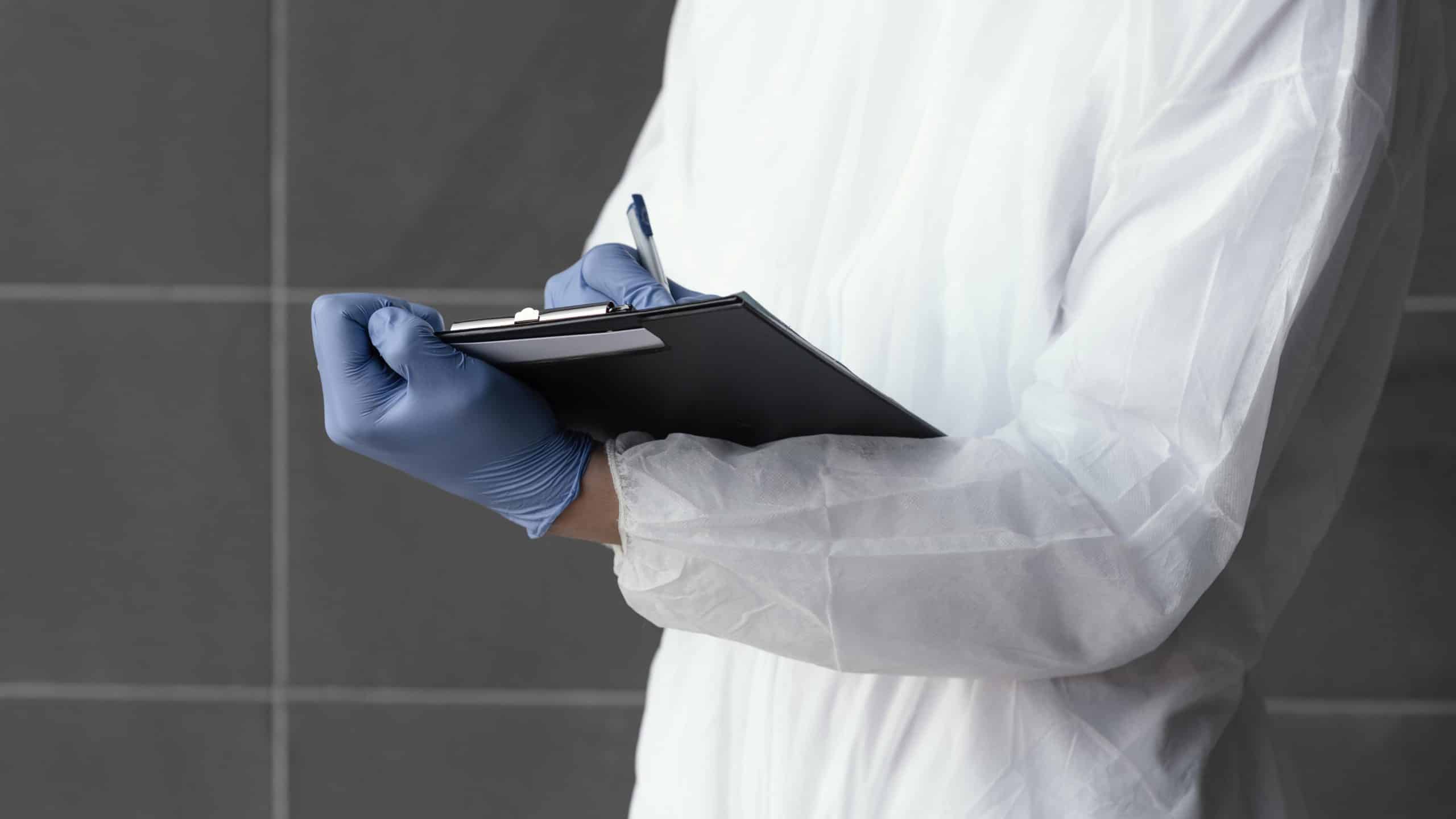 Person in protective gown and blue gloves holds a clipboard and pen, writing or taking notes against a tiled background.