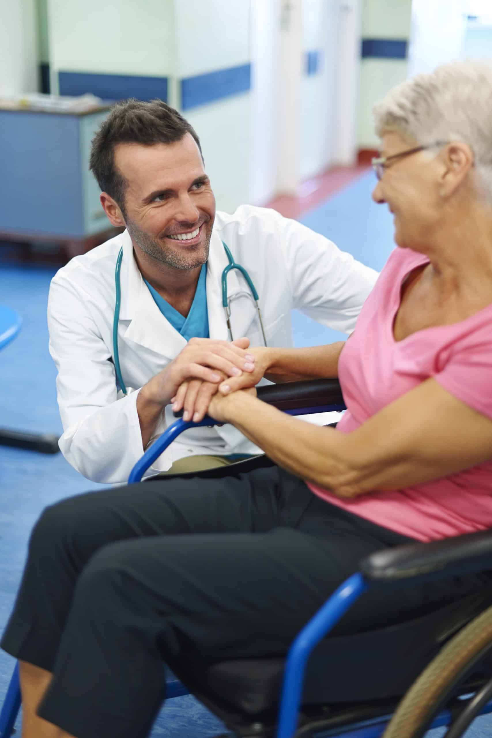 A male doctor kneels beside an older woman in a wheelchair, smiling and holding her hand in a medical facility hallway.