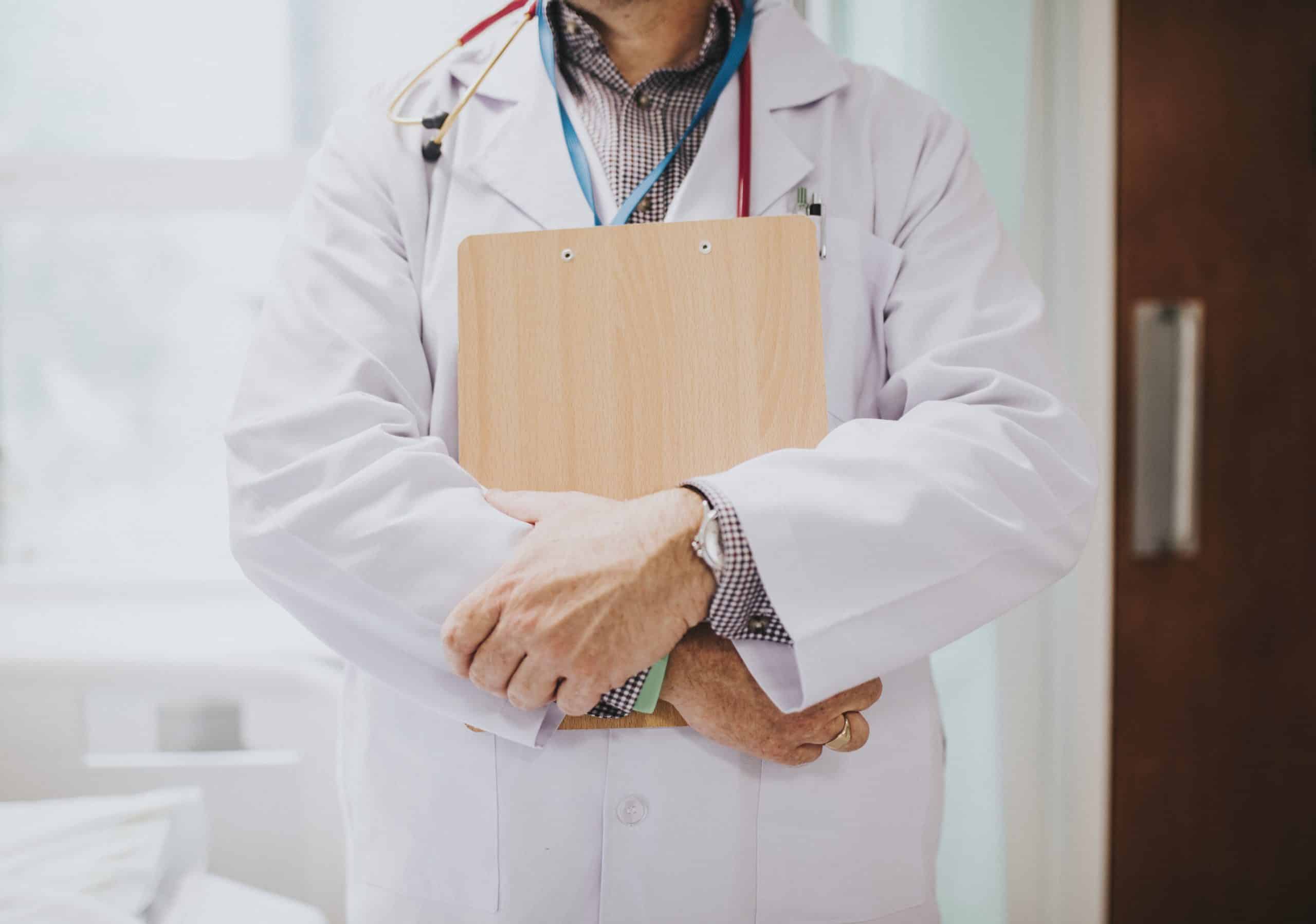 A doctor in a white coat stands with arms crossed, holding a clipboard and wearing a stethoscope around the neck, partially visible in a medical setting.