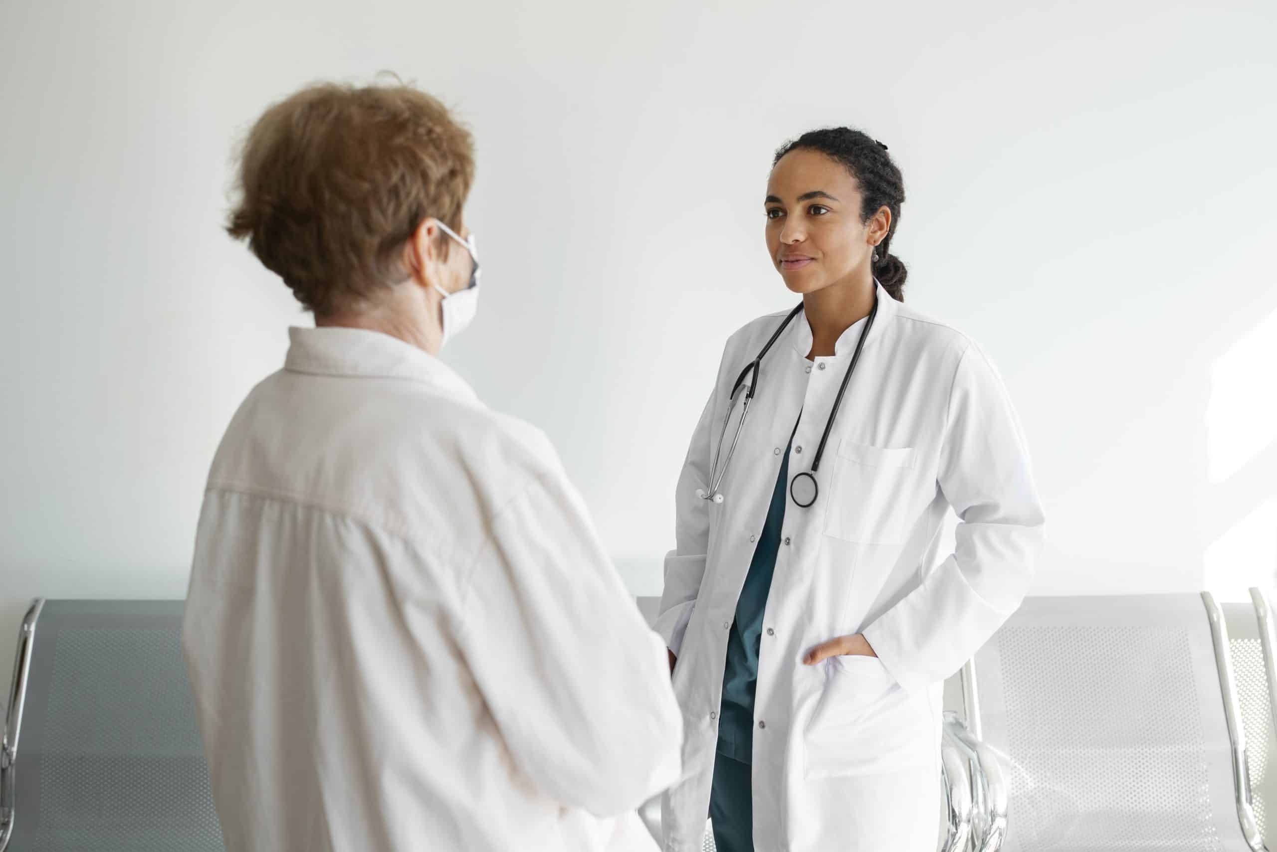 Two healthcare professionals in white coats stand and converse in a clinical setting; one wears a stethoscope and faces the other, who has a mask on.