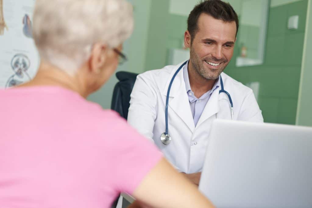A doctor in a white coat with a stethoscope talks to an older woman in a pink shirt while using a laptop in a medical office.
