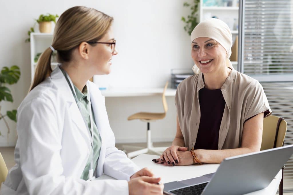 A doctor sits at a desk talking with a patient wearing a headscarf; both are indoors and appear engaged in conversation.