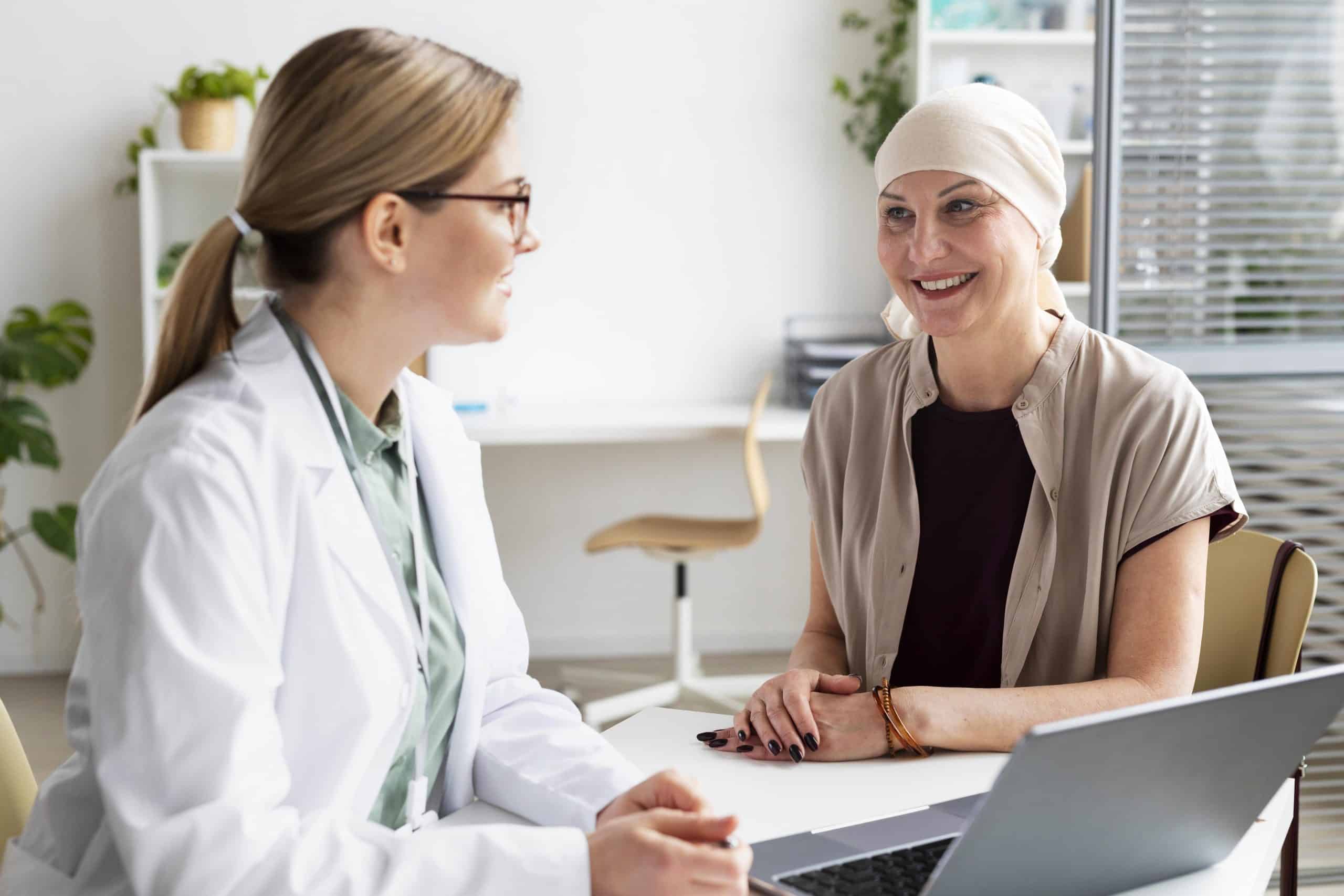 A doctor sits at a desk talking with a patient wearing a headscarf; both are indoors and appear engaged in conversation.