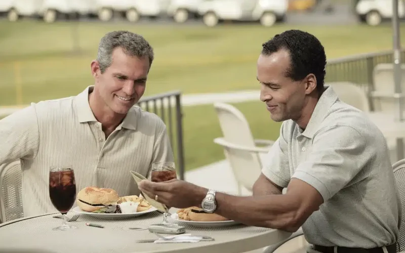 Two men sitting at an outdoor table with food and drinks; one man is paying the bill while the other smiles.