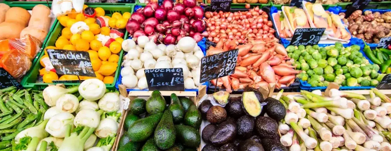 A market stall displays a colorful variety of fresh vegetables, including avocados, onions, garlic, artichokes, leeks, and Brussels sprouts, arranged in neat rows.