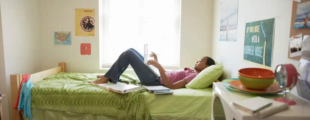 Person lying on a bed in a dorm room, reading a book. The room has a window, posters, books, a microwave, and scattered clothes on the floor.