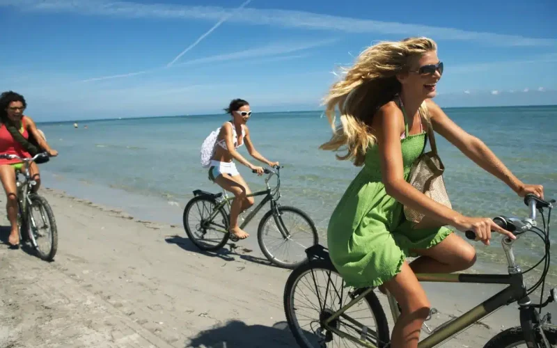 Three people ride bicycles along a sandy beach next to the ocean under a clear blue sky.