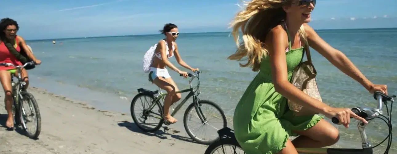 Three people ride bicycles along a sandy beach next to the ocean under a clear blue sky.
