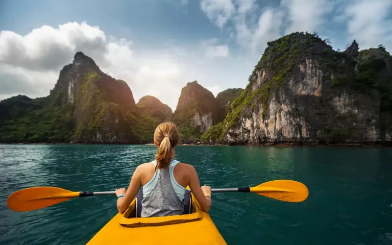 A person kayaks on calm blue water toward large, green, rocky islands under a partly cloudy sky.