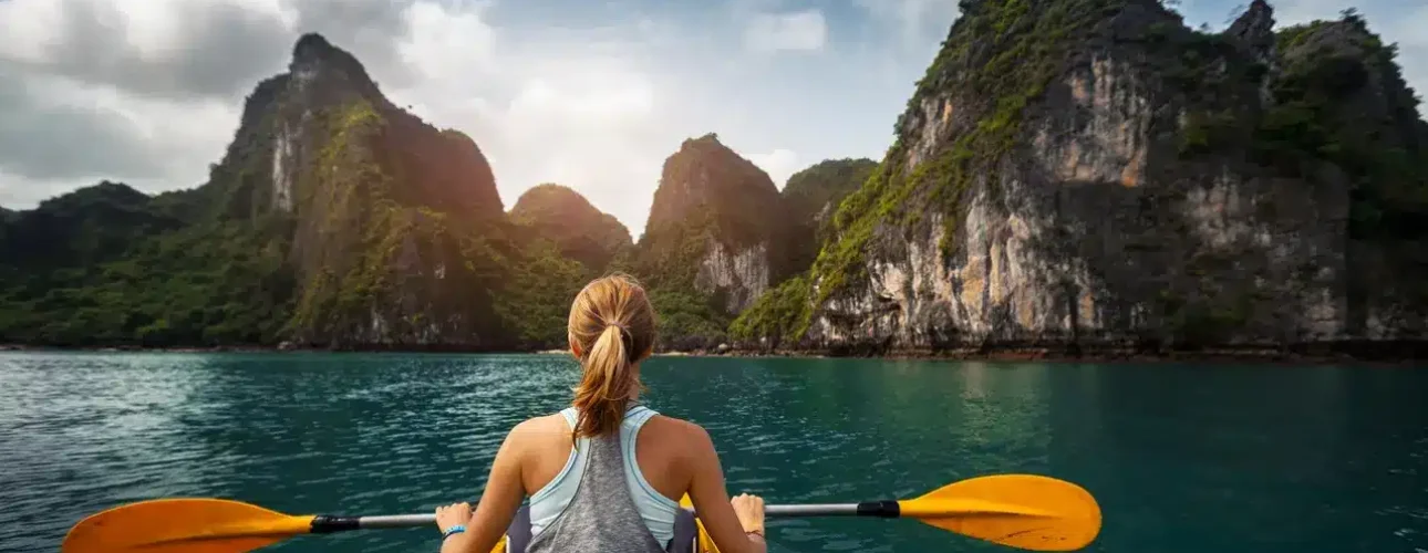 A person kayaks on calm blue water toward large, green, rocky islands under a partly cloudy sky.