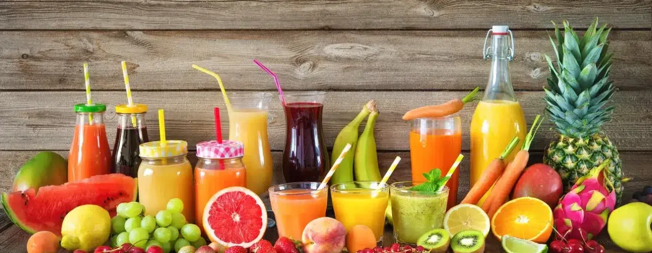 Assorted fresh fruit and vegetable juices in glasses and jars, surrounded by whole and sliced fruits and vegetables on a wooden table.