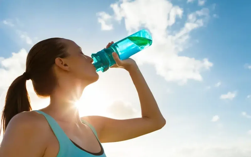 Person in athletic wear drinking water from a bottle outdoors with the sun shining and clouds in the sky.