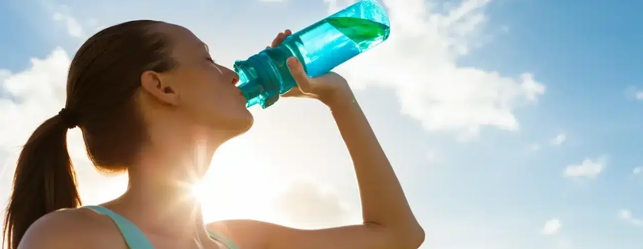 Person in athletic wear drinking water from a bottle outdoors with the sun shining and clouds in the sky.