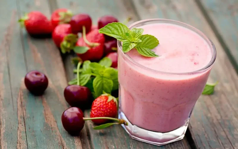 A glass of pink smoothie garnished with mint leaves, placed on a wooden surface next to fresh strawberries, cherries, and additional mint.
