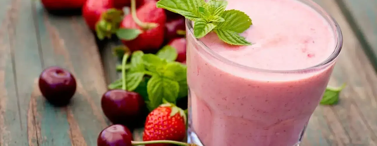 A glass of pink smoothie garnished with mint leaves, placed on a wooden surface next to fresh strawberries, cherries, and additional mint.