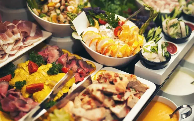 A buffet spread with assorted cold meats, sliced poultry, salads, garnished vegetables, and various dressings arranged on a table.