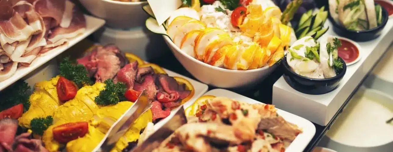 A buffet spread with assorted cold meats, sliced poultry, salads, garnished vegetables, and various dressings arranged on a table.