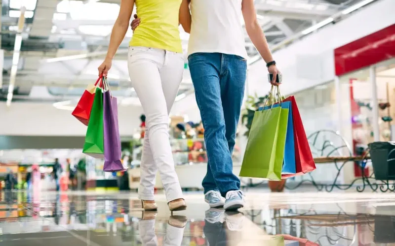 Two people walking in a shopping mall, carrying multiple colorful shopping bags, with their faces not visible in the image.